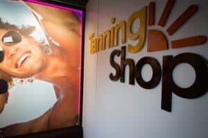 The Tanning Shop 3D gold logo on a salon wall next to a large backlit display image of a laughing tanned man wearing sunglasses