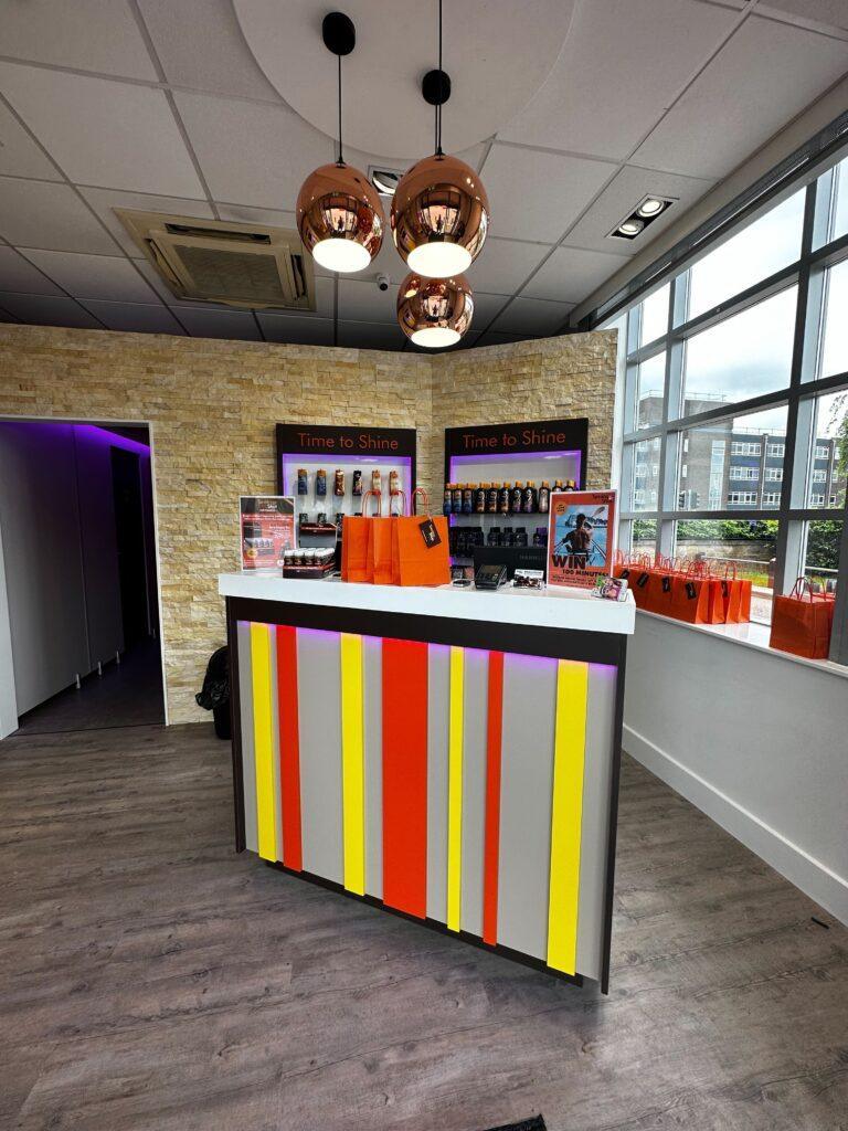 Tanning Shop salon interior showing a colourful red yellow and silver striped reception counter, copper pendant lights, product shelves and Time to Shine signage