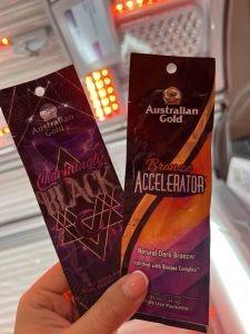Two Australian Gold tanning lotion sachets held up inside a Tanning Shop sunbed room — Absolutely Black and Bronzer Accelerator Natural Dark Bronzer with Banana Complex.