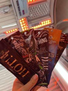 A fan of five Australian Gold tanning lotion sachets held up inside a Tanning Shop sunbed room, including Hot!, Dark, Nothing but Black, and Bronzer Accelerator varieties.
