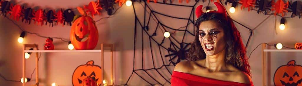 Imgi 26 Halloween Blog Image 2 Woman dressed as a devil with red horns and Halloween makeup surrounded by pumpkins and spider web decorations