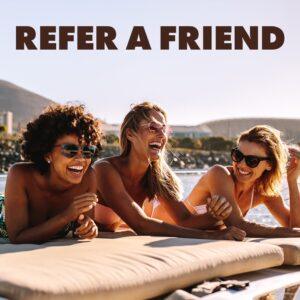 Three laughing women in swimwear and sunglasses lying next to a pool in the sunshine, Refer A Friend text overlay, for The Tanning Shop Refer A Friend page