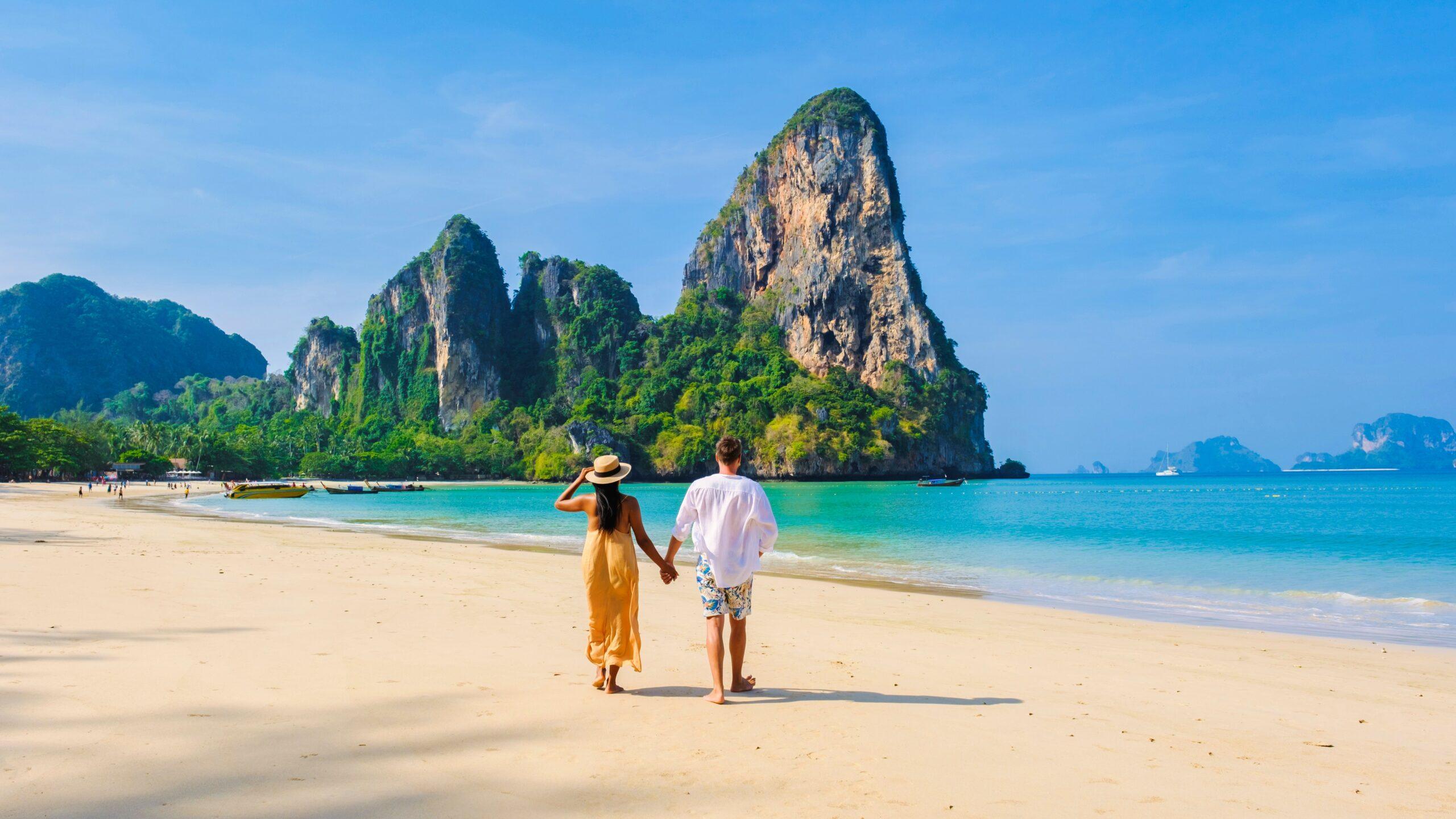 Couple walking hand in hand on Railay Beach in Krabi Thailand with dramatic limestone rock formations and turquoise water, used on The Tanning Shop Our Promises to You page