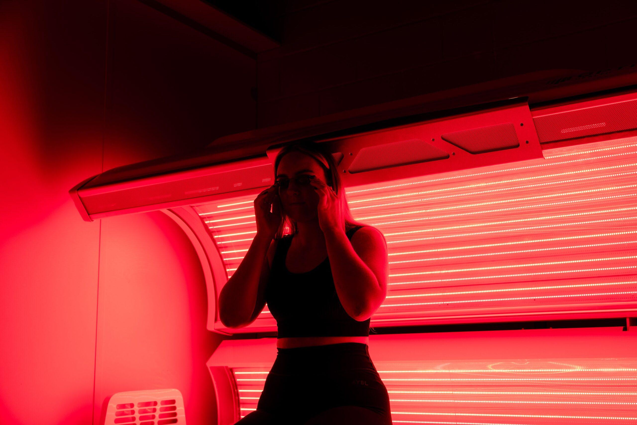 Woman in black activewear standing next to an open red light therapy tanning bed with glowing red LED tubes, used on The Tanning Shop FAQs page