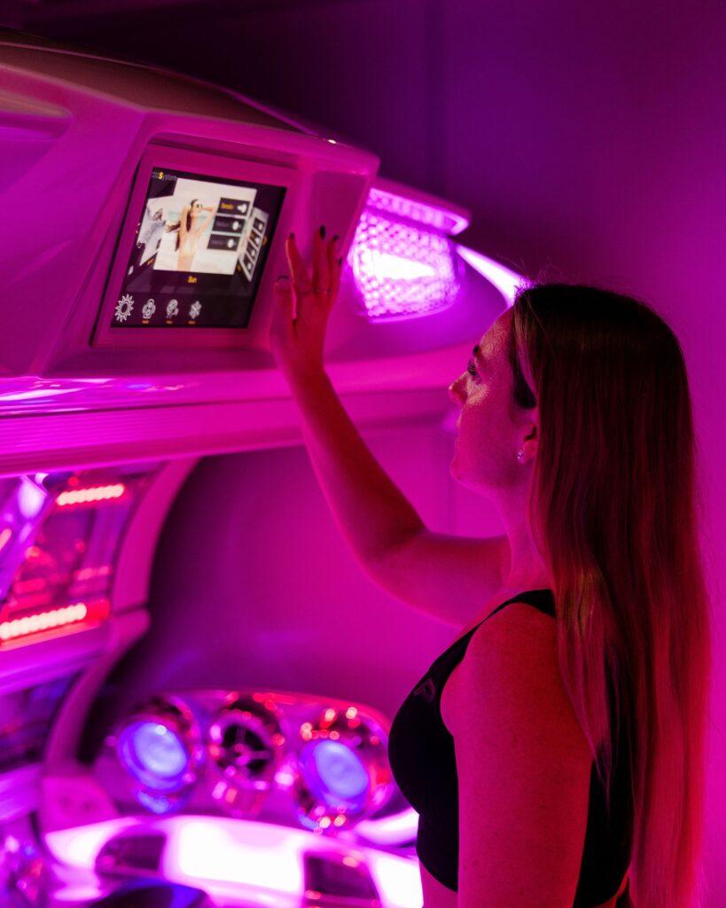 Uv Tanning Shop Woman reaching up to operate the touchscreen control panel of a UV tanning bed in a pink neon-lit Tanning Shop salon room