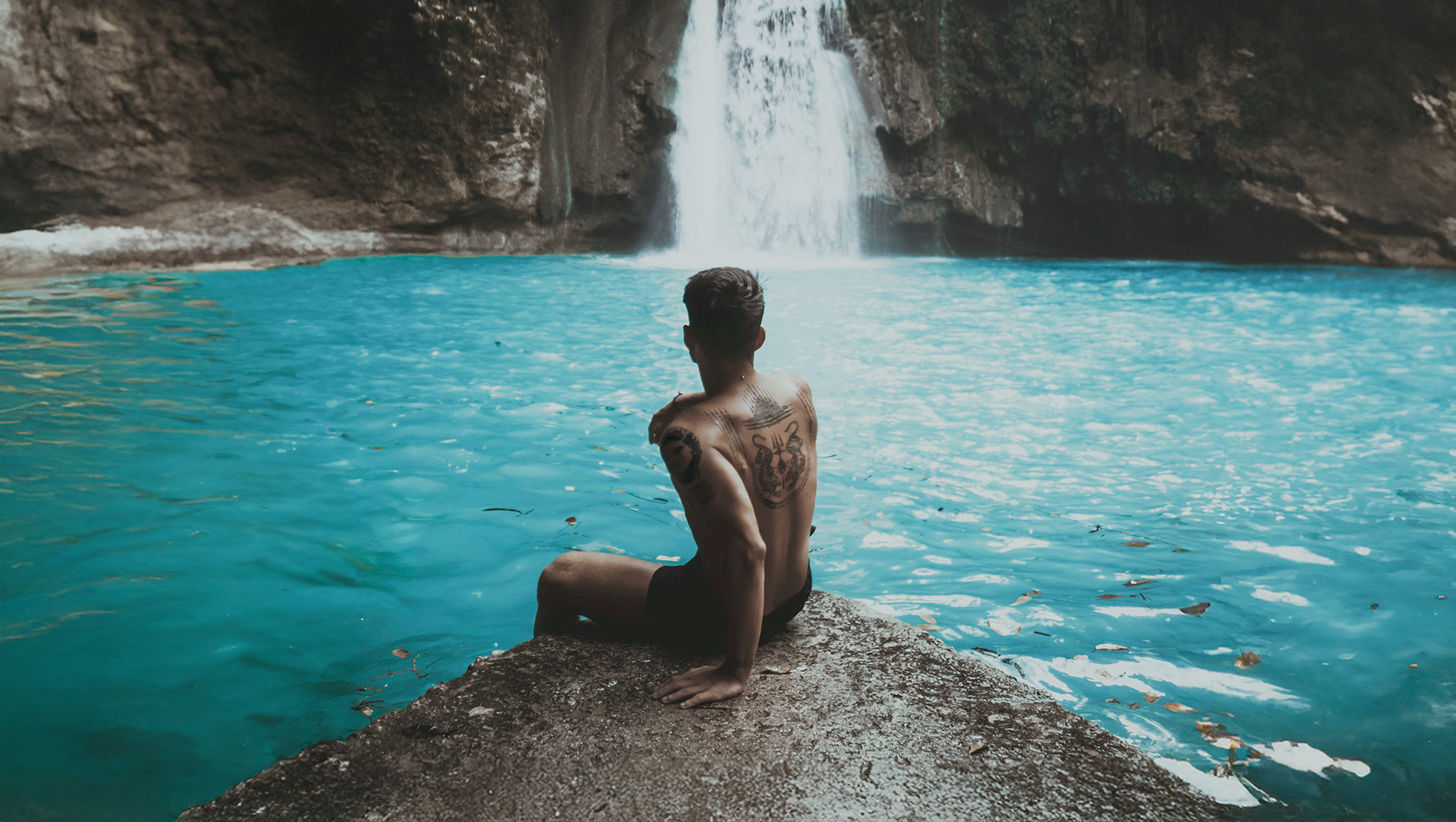Tanned man sitting by a turquoise waterfall pool, representing the lifestyle and content creation opportunities in The Tanning Shop UK influencer programme