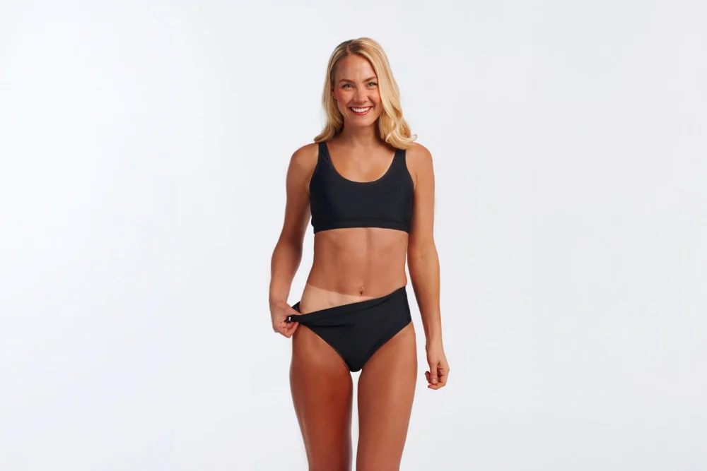 Smiling woman in black swimwear showing a noticeable tan line after getting a Norvell dark spray tan at The Tanning Shop UK.